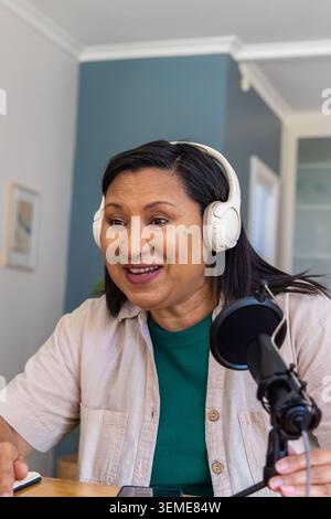 Mature woman speaking into black microphone at home office wearing white headphones and beige shirt Stock Photo