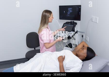 Female tech in scrubs using ultrasound at clinic, African American patient lying on bed, copy space Stock Photo