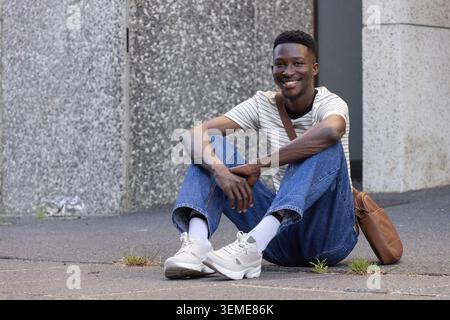 Adult African American man sitting on sidewalk by column, wearing striped tee, holding leather bag Stock Photo
