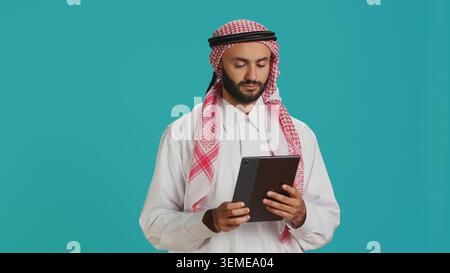 Middle eastern guy browsing tablet on camera, navigating webpages online on digital device. Male model in arabic clothing using gadget to check internet sites, texting on social media. Stock Photo
