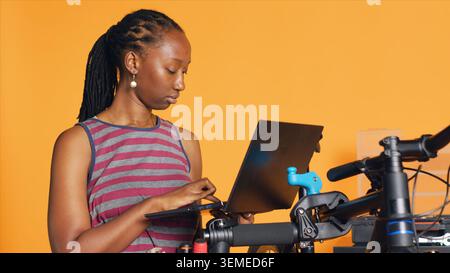 Cheerful mechanic doing bike servicing in studio background atelier shop, looking on laptop maintenance list. Specialist checking online for replacement bicycle parts using notebook, camera A Stock Photo