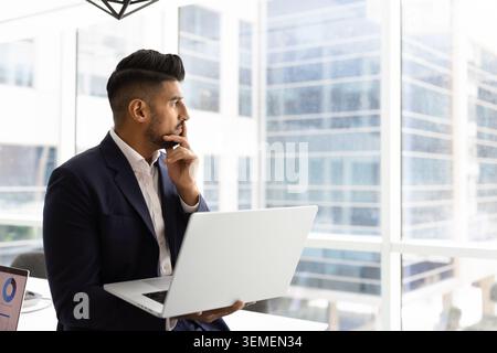 Thoughtful businessman sitting by window with laptop Stock Photo