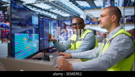 Two Workers Sitting In Warehouse Using Computer For Logistics Management Tasks. Stock Photo
