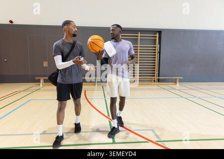 Diverse male friends in sportswear walking and talking on wooden court holding basketball and towel Stock Photo