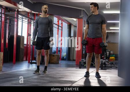 Diverse male training partners holding hex dumbbells in gym wearing gray, red shorts by red rig Stock Photo