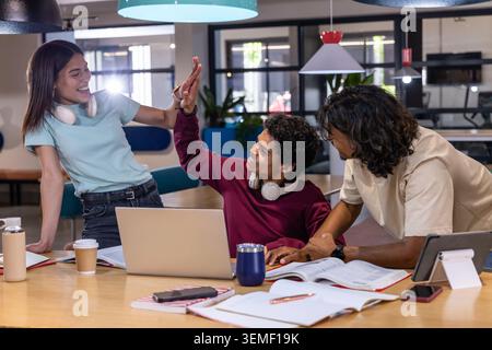 Diverse classmates collaborating around wooden table in communal study, high-fiving at laptop Stock Photo