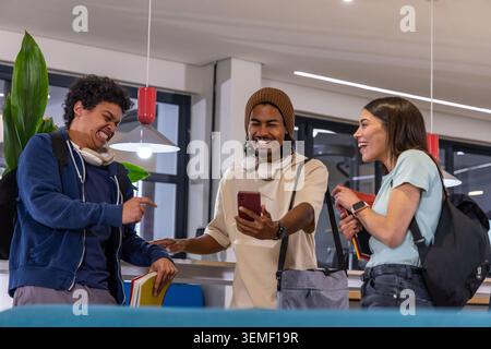 Diverse students laughing, showing smartphone while standing in campus lounge with headphones Stock Photo