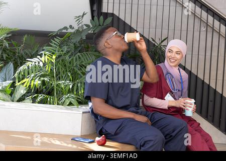 Diverse staff in scrubs with hijab sitting on bench in atrium drinking coffee holding stethoscope Stock Photo