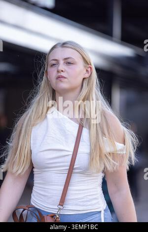 Female in twenties walking past covered path wearing white off-shoulder top and brown crossbody bag. Woman, blonde, urban, transit, canopy, commuter, Stock Photo