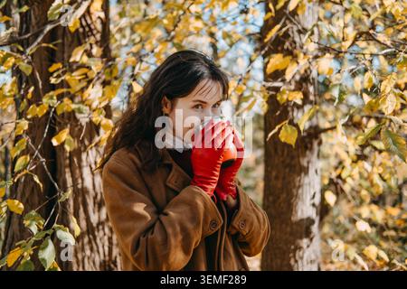 Woman stands in woods holding beverage container with both hands while looking upward. Mindfulness outdoor breathing exercise, positive thinking Stock Photo