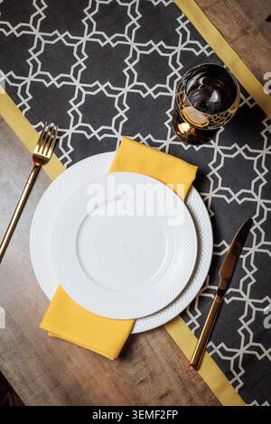 Stylish overhead view of a modern elegant table setting. White textured dinner plates placed on a dark geometric-patterned table runner with gold acce Stock Photo