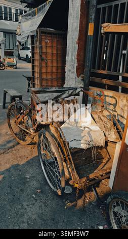Weathered and broken becak (trishaw) with rusted metal frame and worn-out wooden seats, parked on a street corner. Its tires are deflated Stock Photo