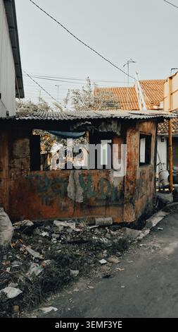 A dilapidated security post with weathered walls covered in faded graffiti stands by a narrow, litter-strewn road. The structure has a corrugated meta Stock Photo