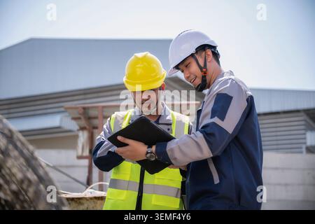 Two construction workers look at tablet together discuss of work. Foreman wears yellow hard hat and high-visibility vest. Engineer wears white hard ha Stock Photo