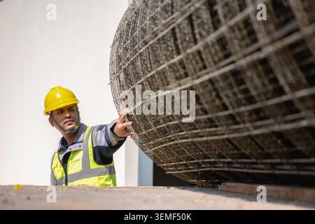 Construction worker wears a yellow hard hat and high-visibility vest. He pushes a large steel mesh coil on concrete. Bright sunlight illuminates the c Stock Photo