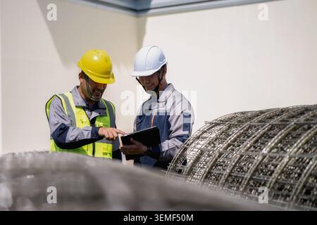 Two construction workers examine a tablet together. One wears a yellow hard hat and high-visibility vest. The other has a white hard hat and gray work Stock Photo