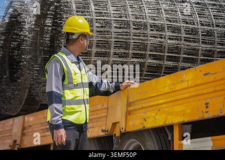 Worker wears a yellow hard hat and safety vest. He checks a large steel coil on a truck. The coil rests on a yellow truck bed. Truck wheels are visibl Stock Photo