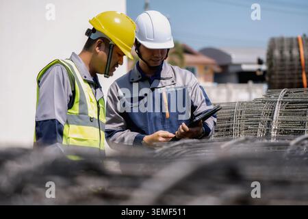 Two construction workers stand together. One wears a yellow hard hat. The other has a white hard hat. Both wear safety vests and work uniforms. One ho Stock Photo