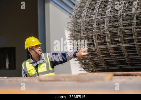 Construction worker pushes large steel mesh roll. He wears yellow hard hat and high-visibility safety vest. Building with windows and concrete backgro Stock Photo