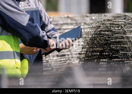 Close up hand of worker in high-visibility vest holds tablet. Another person assists with the device. Large stack of metal pipes dominates the foregro Stock Photo