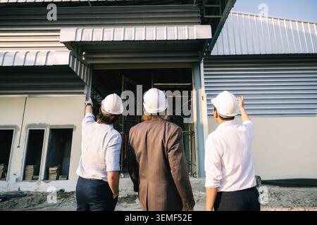 Engineer pointing finger warehouse building structure investor outdoor inspection, Three workers in white hard hats examine a building under construct Stock Photo