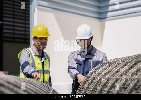 Two construction workers inspect steel reinforcement coils. One wears a yellow hard hat and high-visibility vest. The other sports a white hard hat an Stock Photo