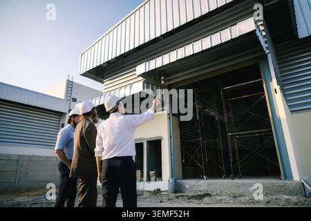 Engineer pointing finger warehouse building structure investor outdoor inspection, Three men discuss a building under construction. Two wear white har Stock Photo