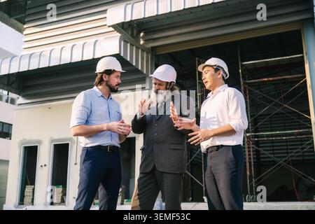 Investor engineers talking outside empty factory warehouse construction site, Three people in white hard hats converse near a building under construct Stock Photo