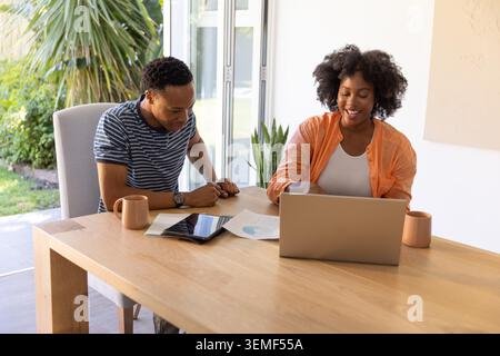 Couple sitting at dining table near sliding glass doors, using silver laptop and tablet with charts Stock Photo