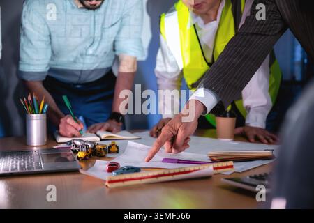close up hand investor pointing construction blueprint boardroom meeting night, Business professionals collaborate around a blueprint. One points to t Stock Photo