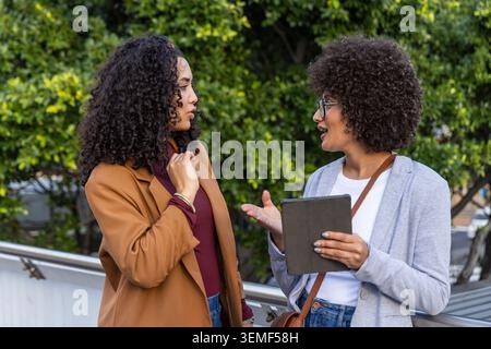 African-American women leaning on rail, talking, holding tablet, wearing camel coat, gray blazer Stock Photo