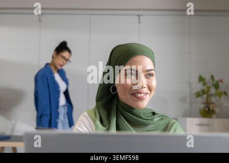 Diverse women team working at desk in office, hijab-wearing woman on laptop, colleague with glasses Stock Photo