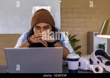 Man wearing brown knit beanie and striped shirt working at home workspace with laptop, VR headset Stock Photo