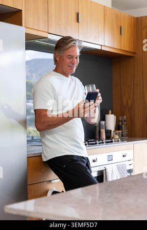 Senior man leaning against counter in modern home kitchen, holding smartphone and glass Stock Photo
