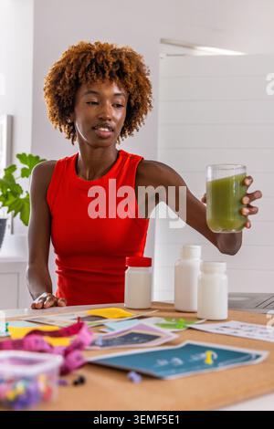 African American woman holding green shake while sorting supplement jars and papers in home kitchen Stock Photo