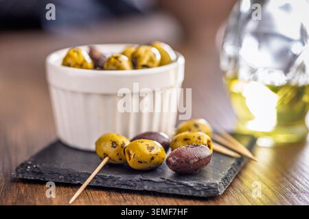 Marinated green and Kalamata olives served in a white ramekin on a slate board with wooden toothpicks, evoking Mediterranean flavors, rustic appetizer Stock Photo