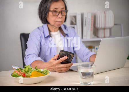 Senior Asian woman using smartphone and laptop while eating fresh salad at home. Elderly lady checking health app, telemedicine or working online with Stock Photo