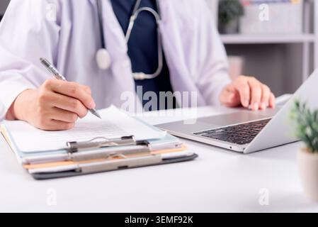 Close up doctor hand writing medical record on clipboard and using laptop computer. Physician working on patient diagnosis report, healthcare insuranc Stock Photo