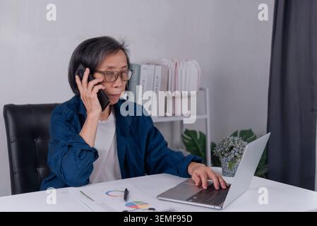 Senior Asian business woman talking on smartphone and working on laptop with financial charts at home office. Elderly entrepreneur multitasking, remot Stock Photo