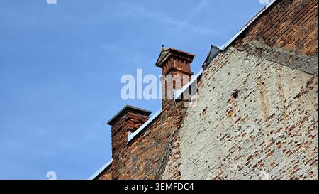 Two weathered brick chimneys extend above the roofline of an old building with crumbling plaster and exposed masonry under a light blue sky Stock Photo