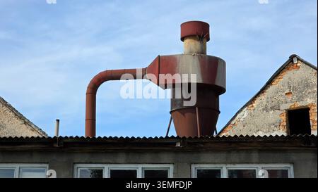 A weathered red metal cyclone separator system sits on a building roof flanked by two triangular gables under a light blue sky with thin clouds Stock Photo
