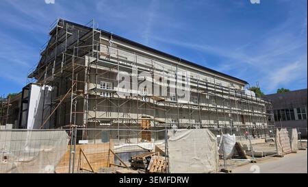 A complex steel scaffolding grid covers the entire length of a gray multi-story building facade behind a mesh perimeter safety fence on a sunny day Stock Photo