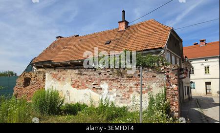 A historic multi-story building features a weathered terracotta tile roof and stands behind a decaying brick and stucco boundary wall overgrown Stock Photo