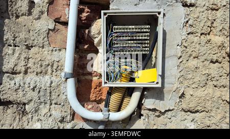 An uncovered plastic junction box displays multiple rows of terminal blocks and a bundle of multi-colored wires connected to conduits on a rough Stock Photo