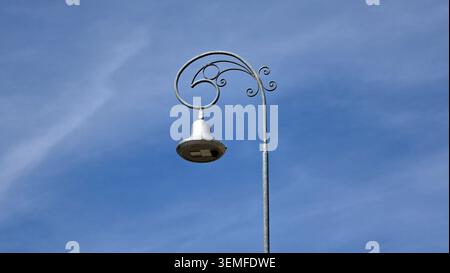 A single street lantern featuring a decorative curved metal bracket and a hanging bell-shaped fixture stands isolated under a blue sky Stock Photo