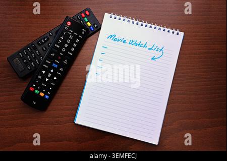 Spiral notebook with "Movie Watch List" and two TV remote controls on a wooden table — planning a watchlist, streaming and entertainment concept. Stock Photo