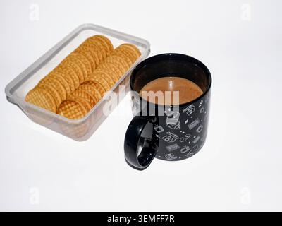 A cup of hot brown milk coffee in a black cup with white retro illustrations, placed next to a clear plastic container containing round biscuits on a bright white background Stock Photo