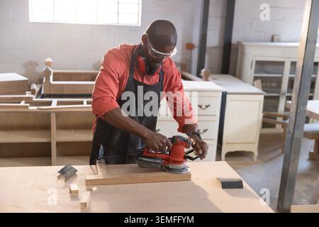 Carpenter sanding wooden plank with red orbital sander at workshop workbench under natural light Stock Photo