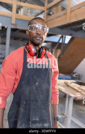 African American man standing in woodshop wearing safety goggles, red earmuffs and black apron Stock Photo