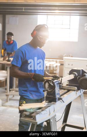African American male carpenters shaping cylinder on lathe in woodshop while colleague using drill Stock Photo